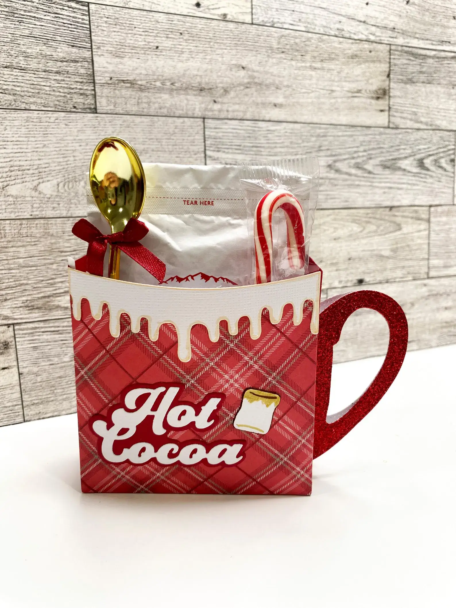 Red plaid mug-shaped gift box filled with hot cocoa mix, a gold spoon, and a candy cane, making a cozy Christmas Eve Box idea for tweens.