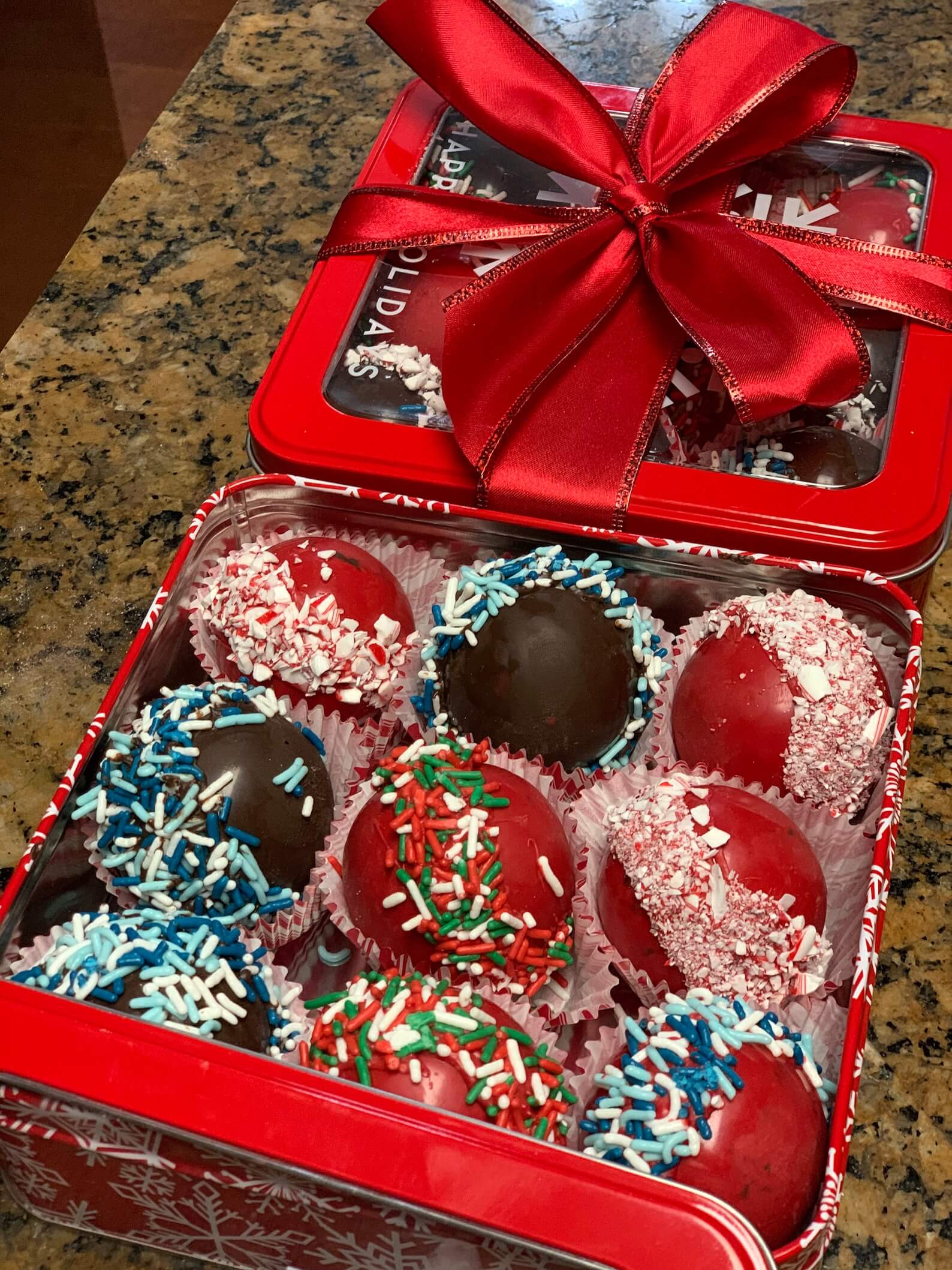Assorted Christmas hot cocoa bombs with peppermint, sprinkle, and chocolate coatings, packed in a red snowflake tin with a satin bow, a sweet addition to any Christmas Eve box for tweens.