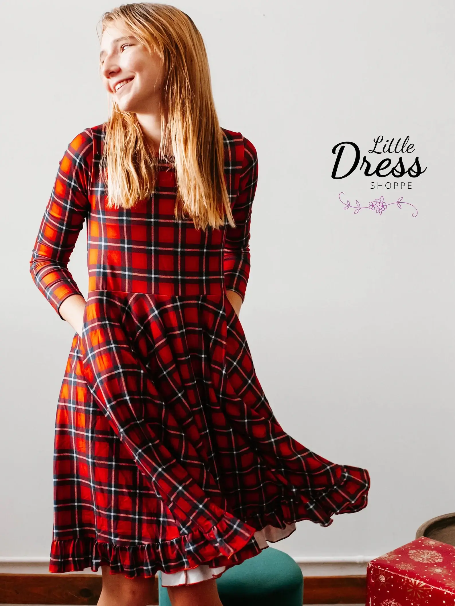 Long sleeve red plaid twirly Christmas dress for tweens with a ruffled hem and pockets, worn by a smiling girl twirling indoors.