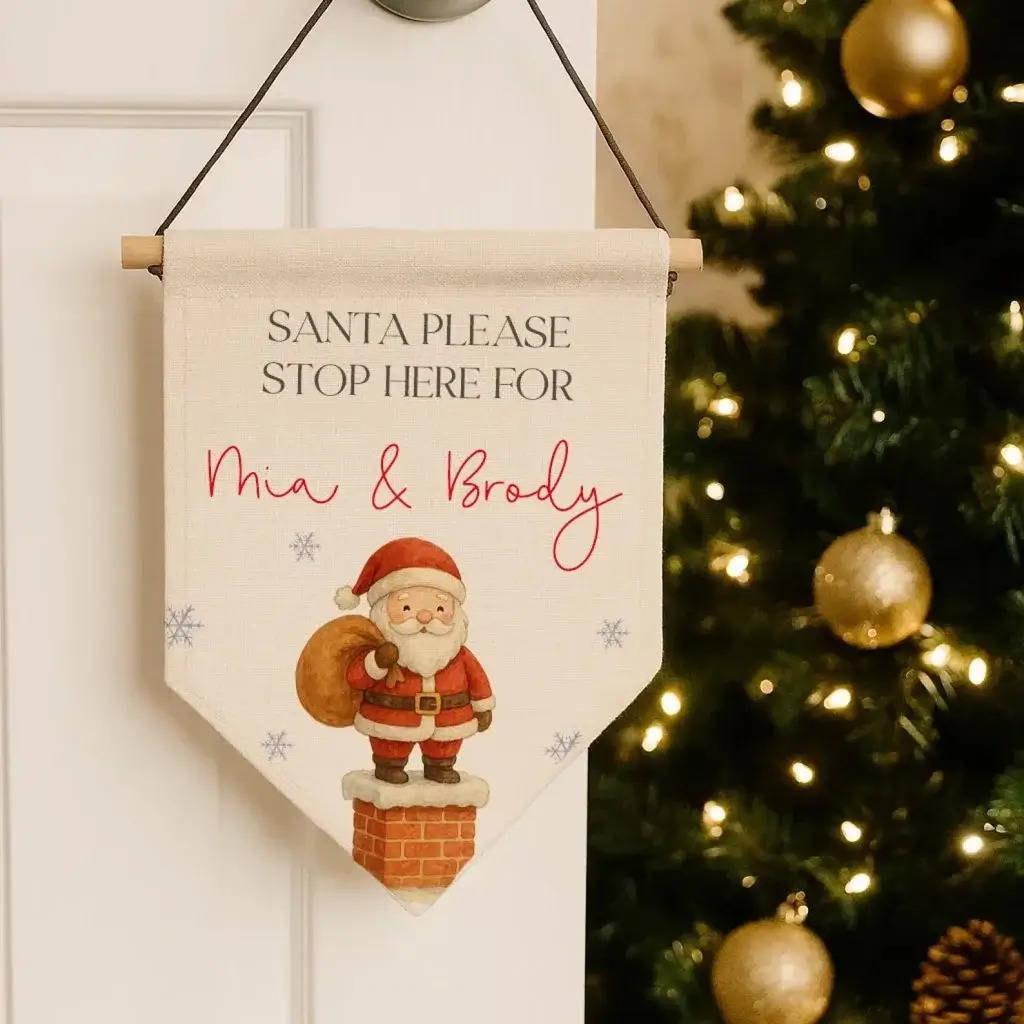 Personalized "Santa Please Stop Here" fabric flag with custom names and Santa illustration, hung by a Christmas tree as a festive door decoration.