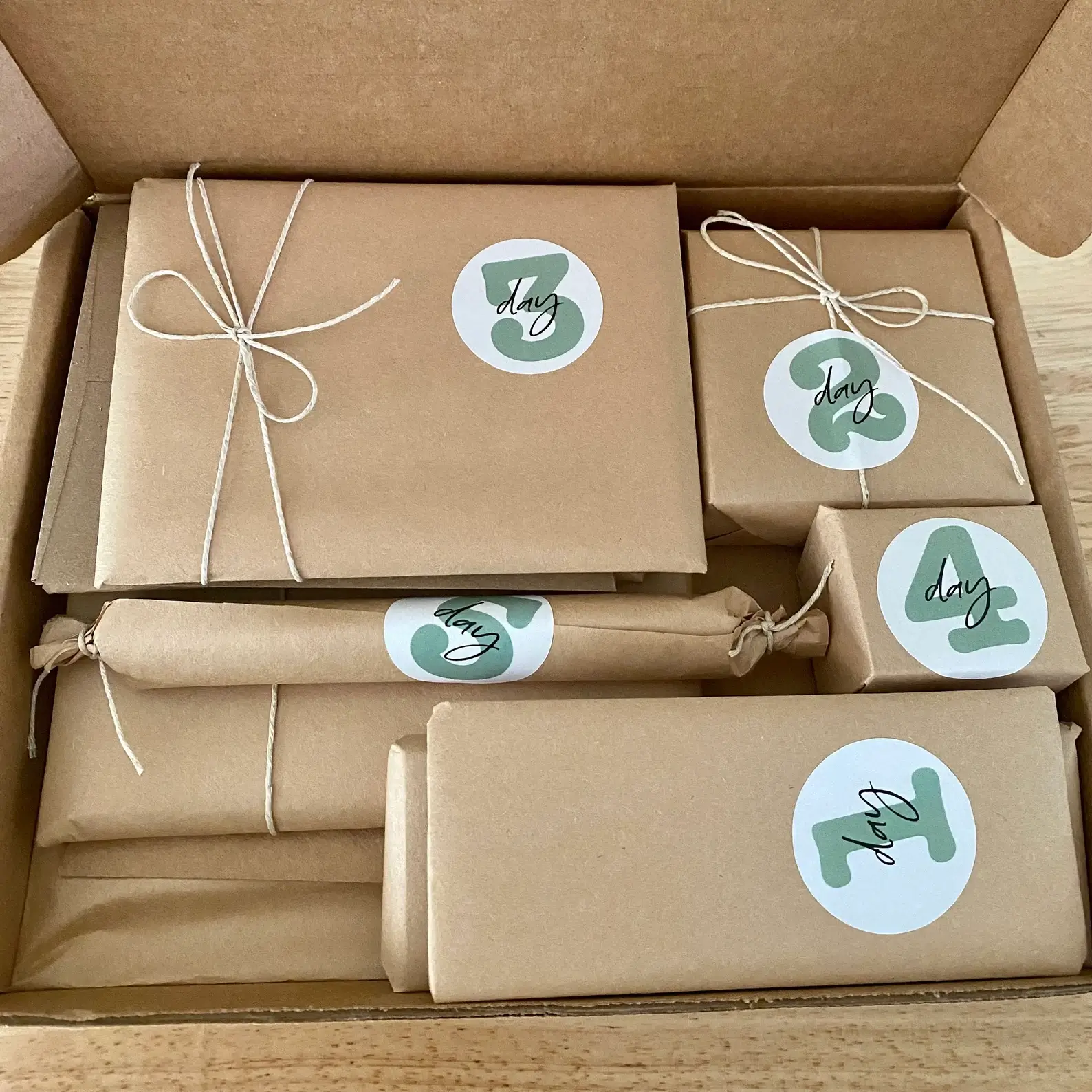 Tween advent calendar with kraft-wrapped gifts numbered "day 1" through "day 4" in sage-green stickers, tied with twine inside an open gift box.